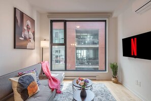A modern living room with a sofa, a coffee table with flowers, a TV, and a large window with a view of a building. - 1 BR Old Port| Basilica (Montreal)
