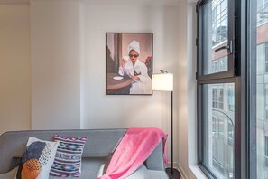 A modern hotel room with a grey sofa, a pink throw blanket, a framed artwork, a floor lamp, and a large window with a city view. - 1 BR Old Port| Basilica (Montreal)