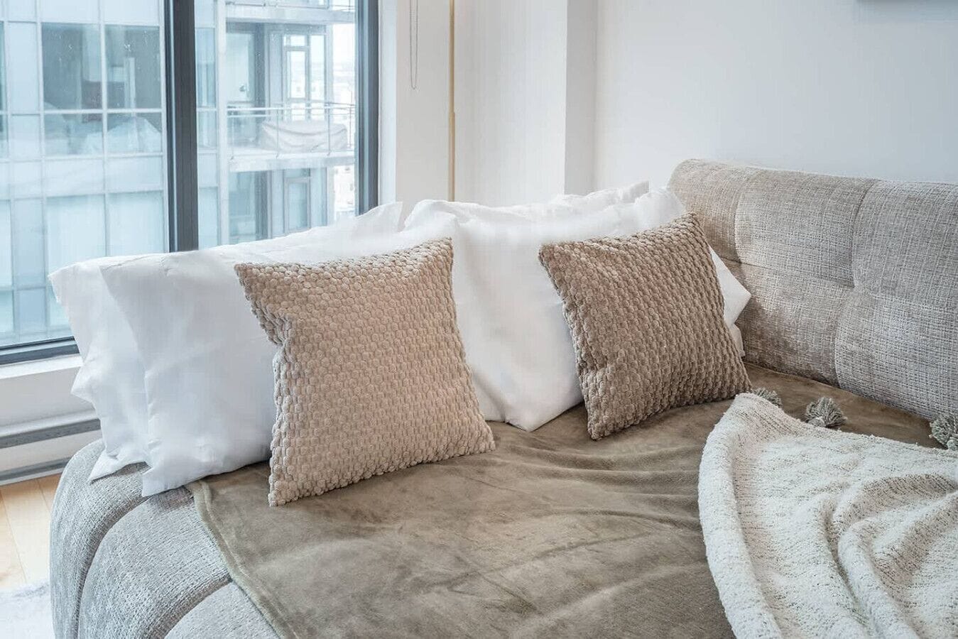 Un salon moderne avec un canapé gris, des coussins blancs à texture intéressante et une vue sur le paysage urbain, visible à travers une grande fenêtre.