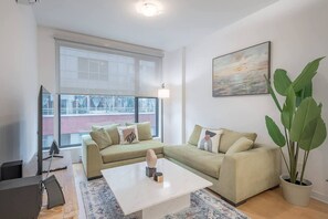A modern living room with a sectional sofa, a coffee table, a TV, and a large window with a view of buildings. - 1 BR Old Port| Basilica (Montreal)