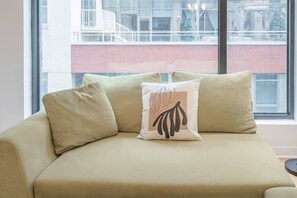 Um sofá bege com três almofadas, uma delas com um desenho de folha, em um quarto com uma janela grande que dá para um edifício de fachada em tijolos vermelhos.