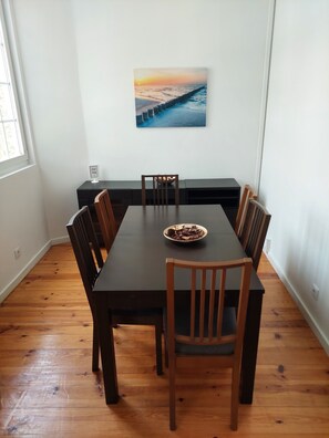 A dining area with a long table, wooden chairs, and a painting of a sunset over the ocean. - Lightoasis Oeiras 2 (Oeiras)