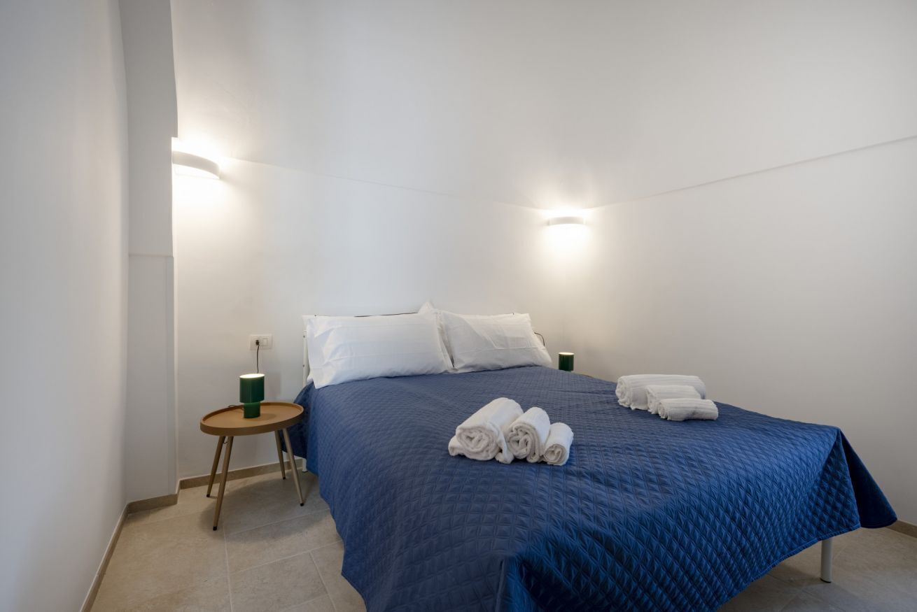 A single bed with a blue quilt, white pillows, and rolled towels on a white wall with two wall-mounted lights.