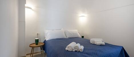 A single bed with a blue quilt, white pillows, and rolled towels on a white wall with two wall-mounted lights.