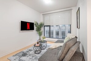 Ein modernes Wohnzimmer mit einem grauen Sofa, einem an der Wand befestigten Fernseher, einer Topfpflanze und einem großen Fenster.