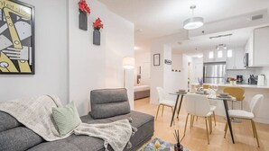 Ein modernes Wohnzimmer mit einer grauen Couch, einem Essbereich mit weißen Stühlen und einer Küche mit weißen Schränken.