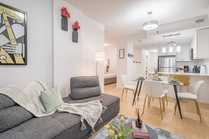 Ein modernes Wohnzimmer mit einer grauen Couch, einem Essbereich mit weißen Stühlen und einer Küche mit weißen Schränken.