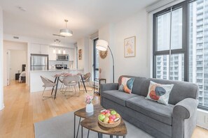 Un apartamento moderno con sofá gris, comedor, cocina y ventanas amplias.
