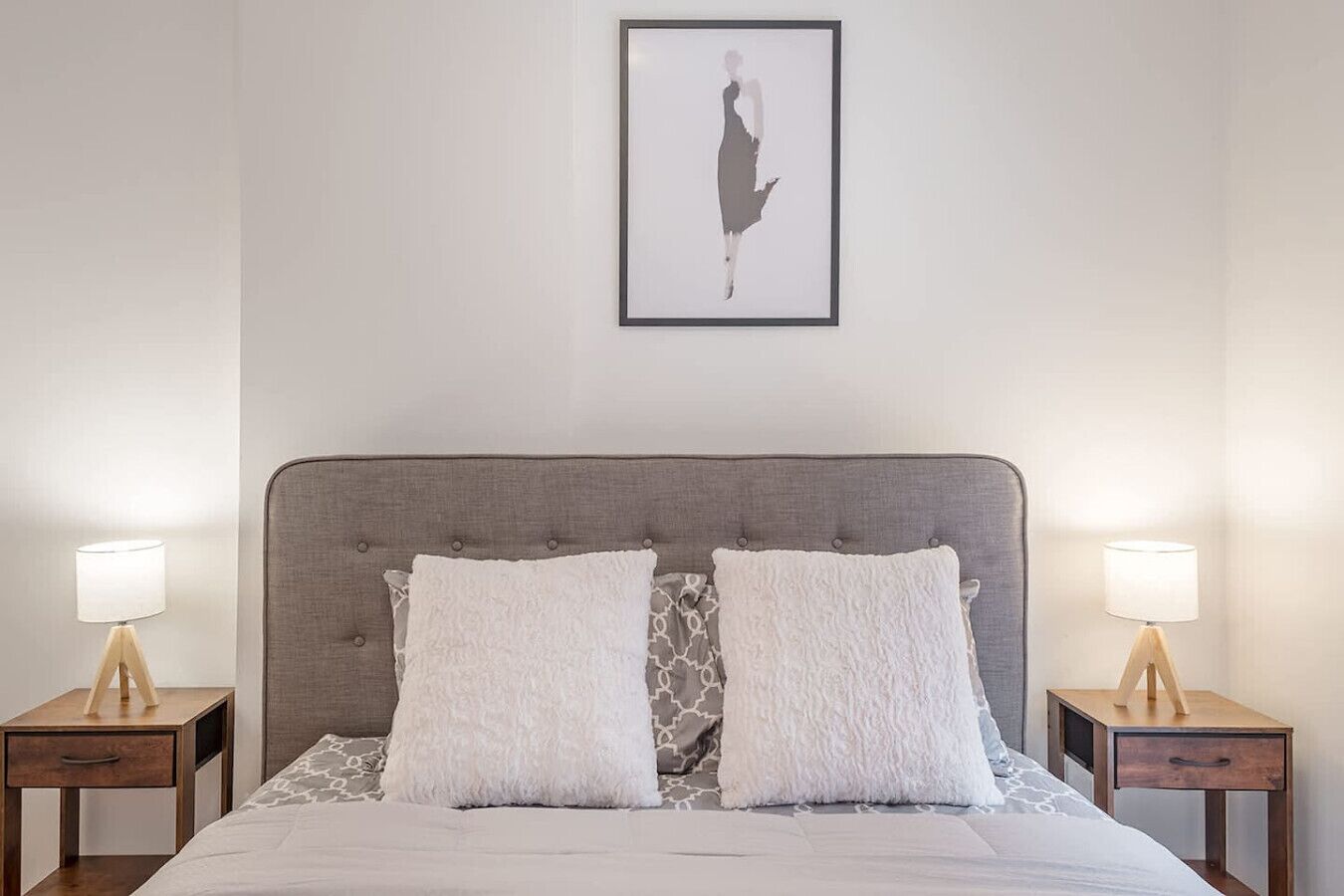 A bedroom with a grey upholstered headboard, two bedside tables with lamps, and a framed artwork above the bed.