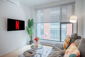 Un soggiorno moderno con una grande finestra, una TV a muro che mostra il logo di Netflix, una pianta in vaso, un tavolino con un vaso di fiori e un divano con cuscini colorati.