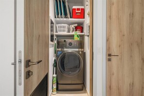 A compact laundry room with a front-loading washing machine, a drying rack, and storage shelves. - Close to Eagle Beach·Balcony·Pool & Jacuzzi·BBQ & (Eagle)