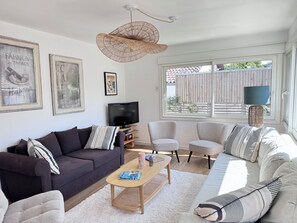 Ein Wohnzimmer mit einem dunklen Sofa, hellen Sesseln, einem Holztisch und einem großen Fenster mit Blick auf einen Holzzaun.