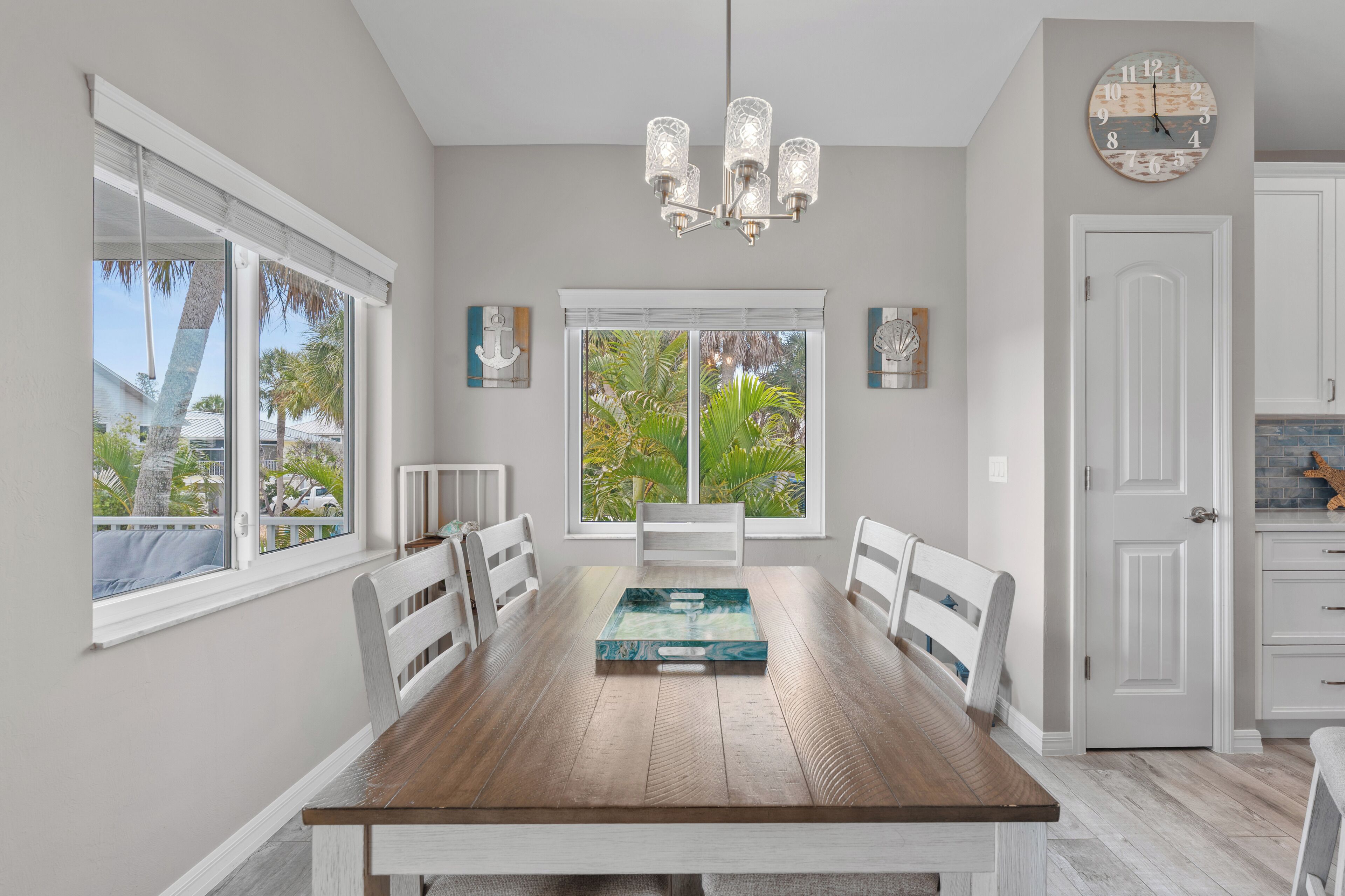 A dining room with a wooden table, white chairs, a chandelier, and a view of palm trees through large windows.