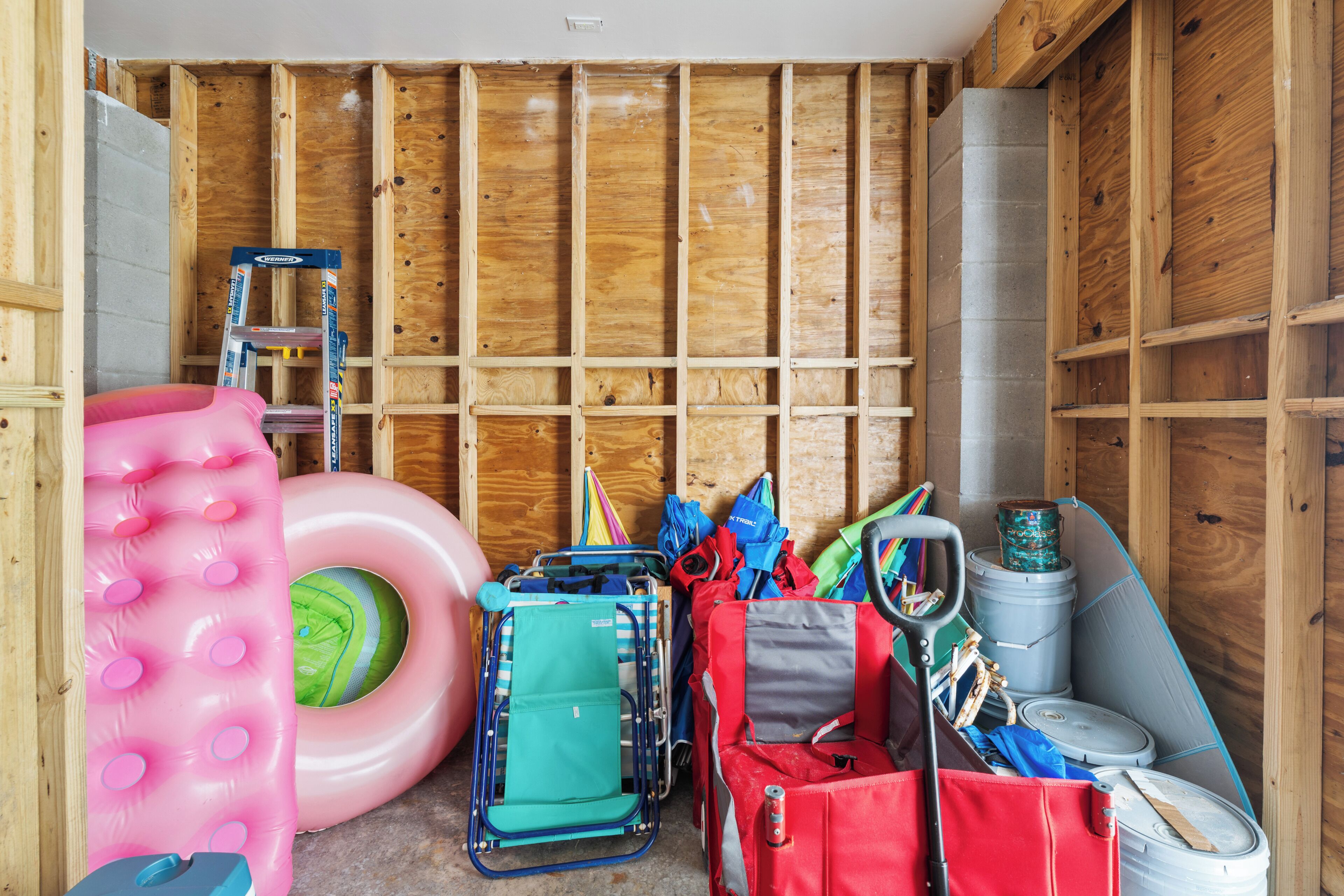 A room with wooden walls, a ladder, a pink inflatable pool, a green inflatable pool, a blue folding chair, a red bag, and a gray bucket.