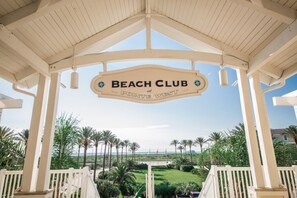 L’entrée d’un club de plage, avec un panneau indicateur, des palmiers et une vue sur la plage.