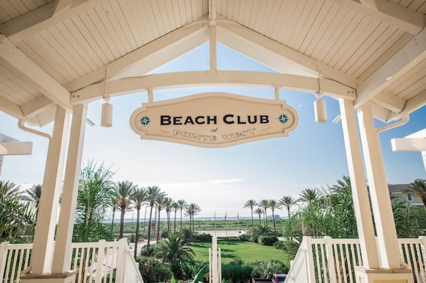 A beach club entrance with a sign, palm trees, and a view of the beach.