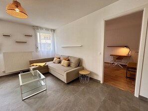 Una sala moderna con un sofá, una mesa de centro de vidrio, una mesita auxiliar, un radiador, una ventana con cortinas y un escritorio con una silla.