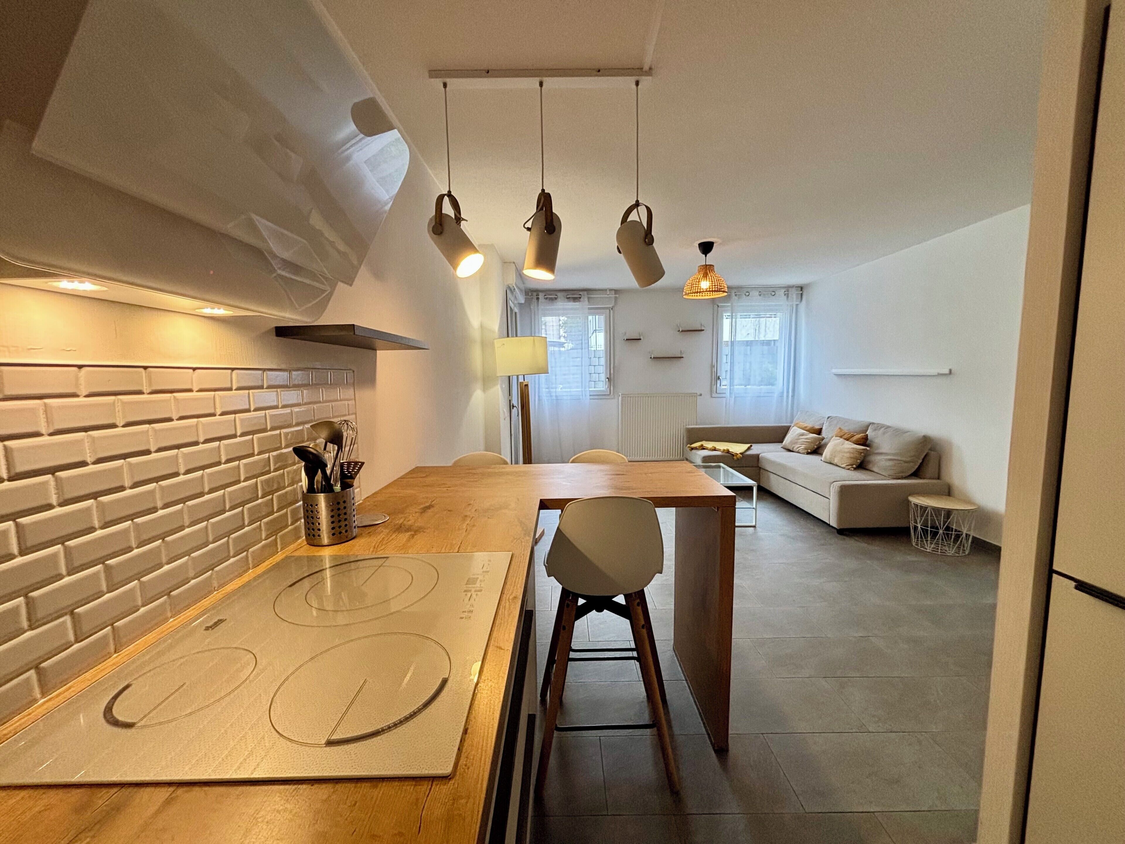 Une cuisine moderne avec un mur de fond en briques, un plan de travail en bois et une cuisinière intégrée. On y trouve un coin repas avec une table et des chaises, ainsi qu’un espace de vie avec un canapé et une petite table d’appoint.