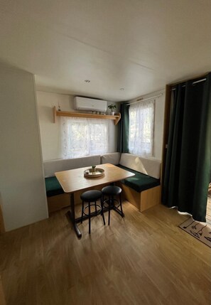 Quarto pequeno com uma mesa de madeira, dois bancos pretos, um banco, uma prateleira pequena com uma planta e uma janela com cortinas leves.