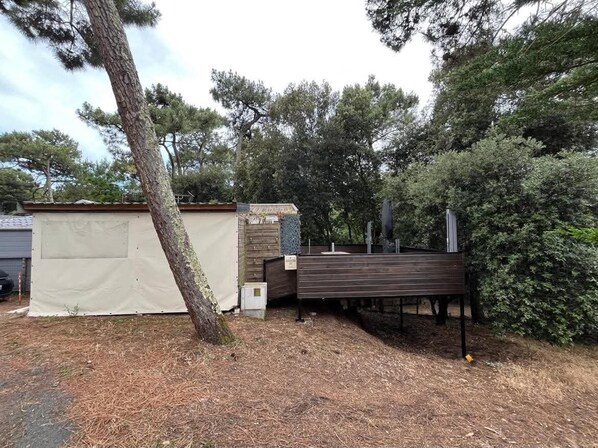 Un espacio al aire libre cubierto con una terraza de madera, un pequeño edificio con techo inclinado y árboles densos alrededor.