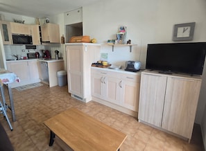 A compact kitchen with a microwave, toaster, and refrigerator. There's a wooden table and a bench in the center.