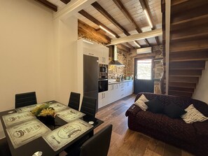 A dining area with a table set for two, a kitchen in the background, and a dark-colored sofa with patterned cushions. - YourHouse A Casa Do Cordeiro (Allariz)