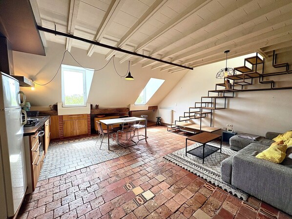 Eine Loft-artige Wohnung mit Küche, Essbereich und Wohnbereich, die freiliegende Holzbalken, einen Backsteinboden und eine Treppe mit geometrischen Regalen zeigt.
