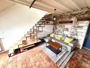 Eine Treppe mit Holzdielen und Metallgeländern, ein graues Sofa mit gelben Kissen, ein Holztisch und ein gemustertes Teppich auf einem Backsteinboden.