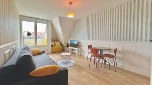A modern living room with a sofa, a dining table, and chairs, a TV, and a large window. - Studio with balcony in Villers-sur-Mer (Villers-sur-Mer)