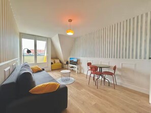 A modern living room with a sofa, a dining table, and chairs, a TV, and a large window. - Studio with balcony in Villers-sur-Mer (Villers-sur-Mer)