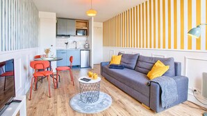 A living room with a grey sofa, red chairs, a round coffee table, and a kitchen in the background. - Studio with balcony in Villers-sur-Mer (Villers-sur-Mer)
