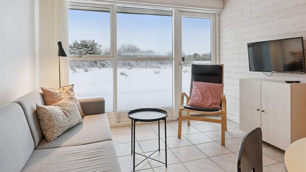 Una sala de estar moderna con un sofá, una silla, una mesita, un televisor y vistas a un paisaje nevado a través de amplias ventanas.