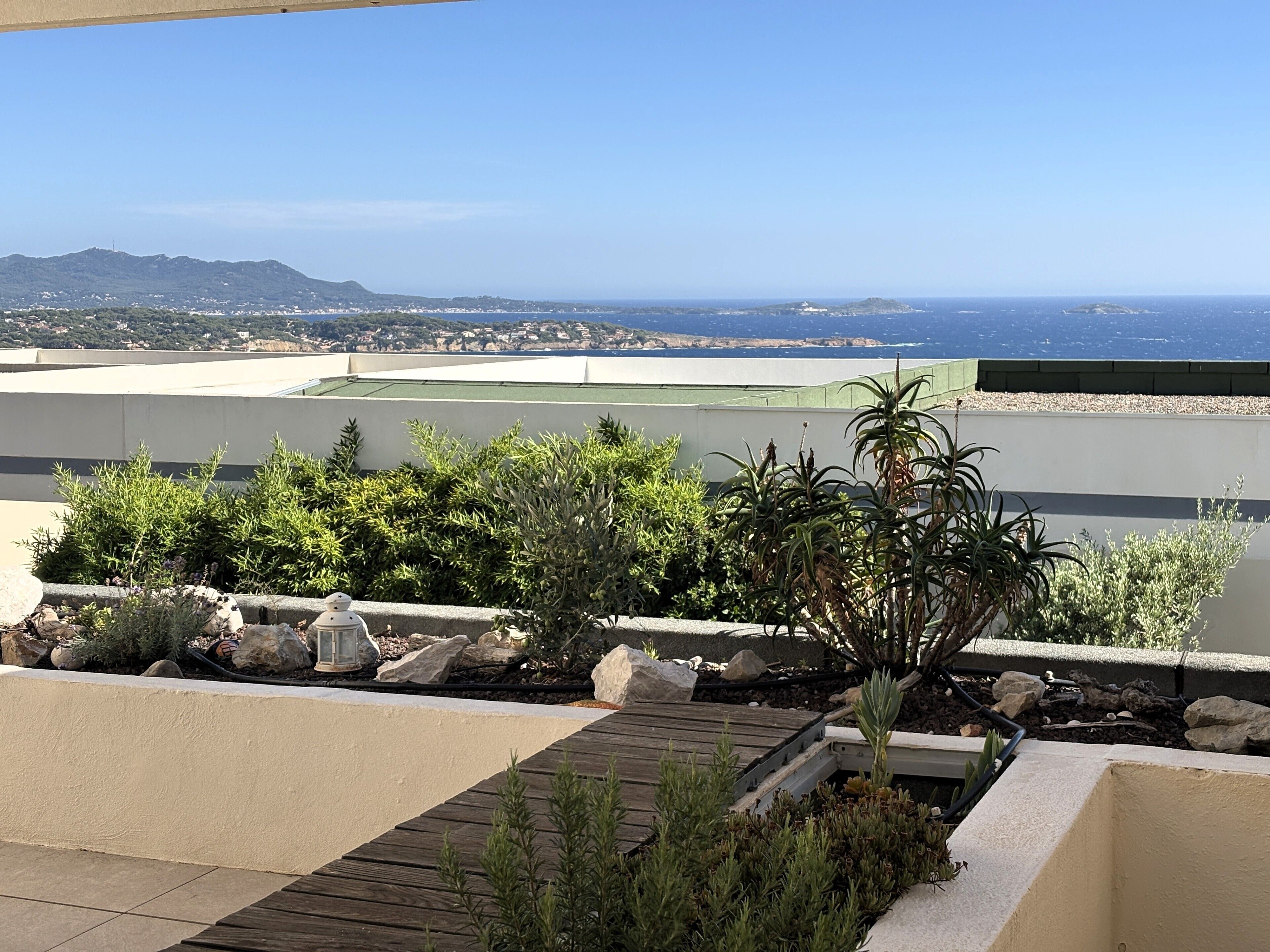 A rooftop terrace with a view of the ocean and distant hills, featuring a wooden deck and various plants.