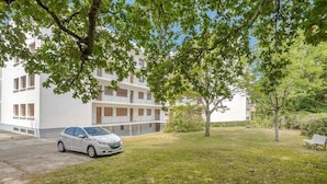 A white building with a car parked in front, surrounded by trees and grass. - Apartment with balcony and parking near the beach (Arcachon)