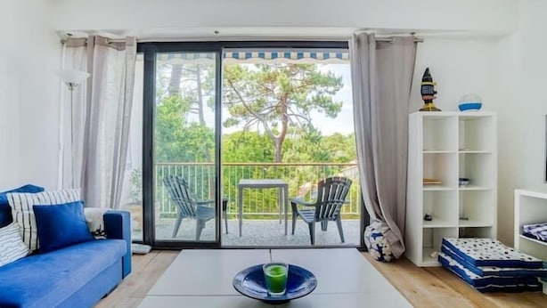A room with a blue sofa, a white coffee table with a green glass, a balcony with chairs, and a white shelving unit. - Apartment with balcony and parking near the beach (Arcachon)