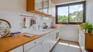 A modern kitchen with white cabinets, a wooden countertop, a window with a view, and a hanging rack with kitchen utensils. - Apartment with balcony and parking near the beach (Arcachon)