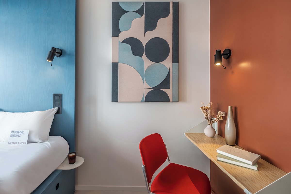 A hotel room with a bed, a desk with a chair, a wall art piece, and a vase with dried flowers.
