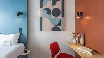 Une chambre d’hôtel comprenant un lit, un bureau avec une chaise, une œuvre d’art murale et un vase avec des fleurs séchées.