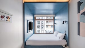 Un dormitorio con cama, ventana, televisión instalada en la pared y una mesita.