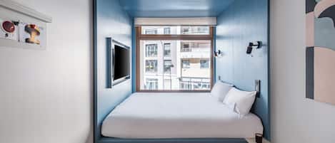 Une chambre à coucher avec un lit, une fenêtre, un téléviseur fixé au mur et une petite table.