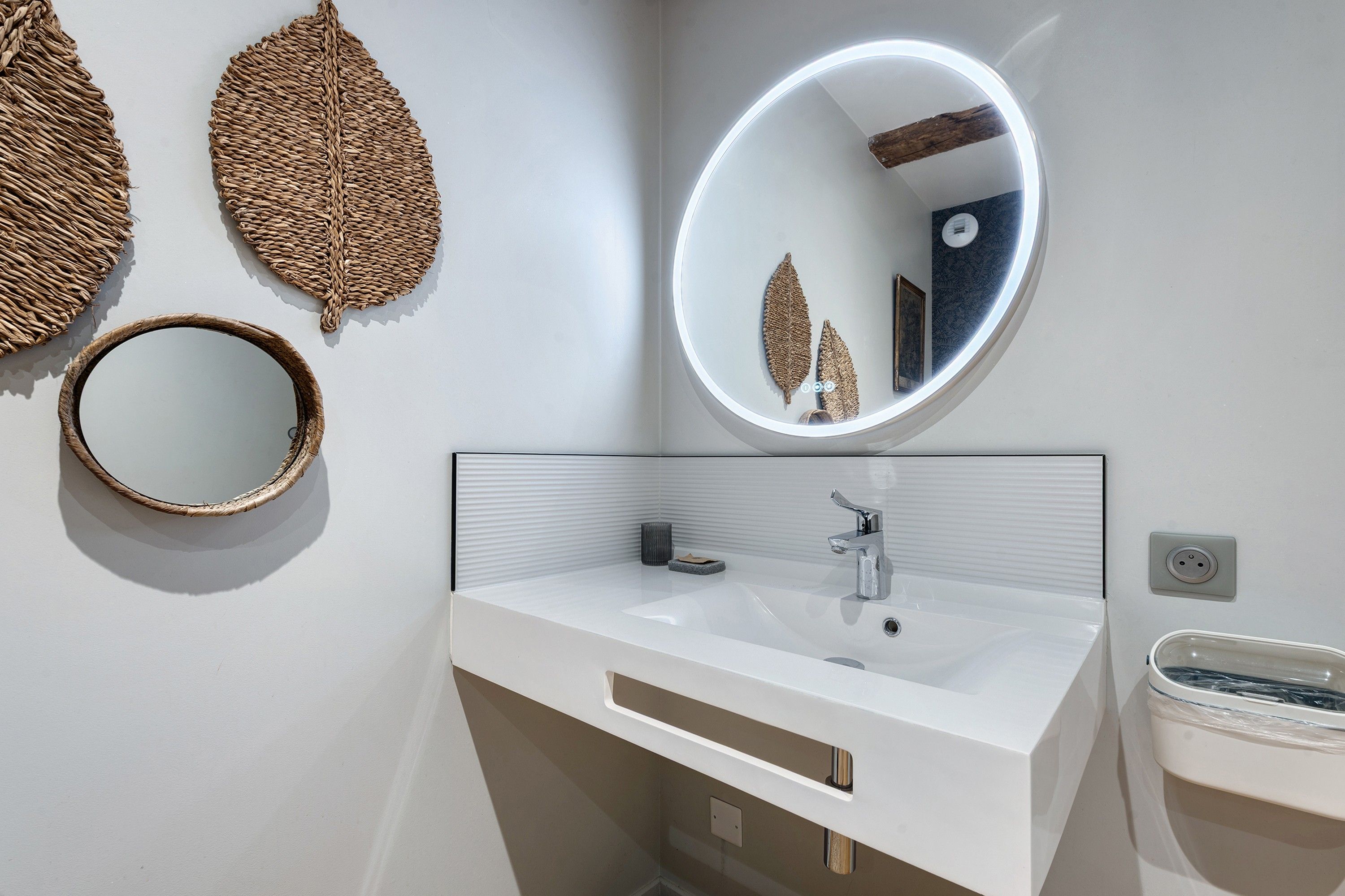 Une salle de bain avec un meuble-lavabo blanc, un miroir rond et deux feuilles décoratives en forme de motifs tissés accrochées au mur.