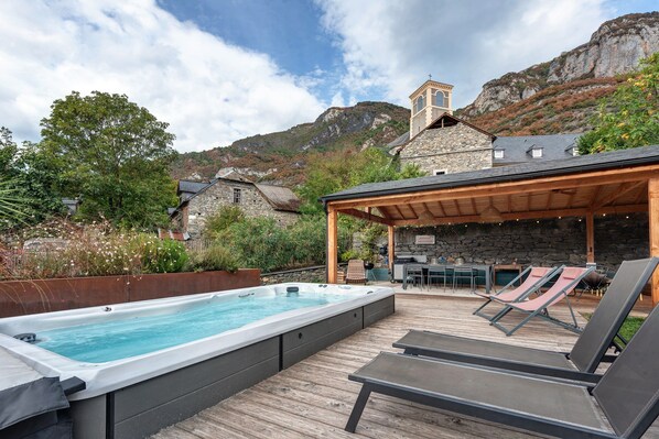 A hot tub on a wooden deck with lounge chairs, a stone building with a bell tower, and a mountainous backdrop.