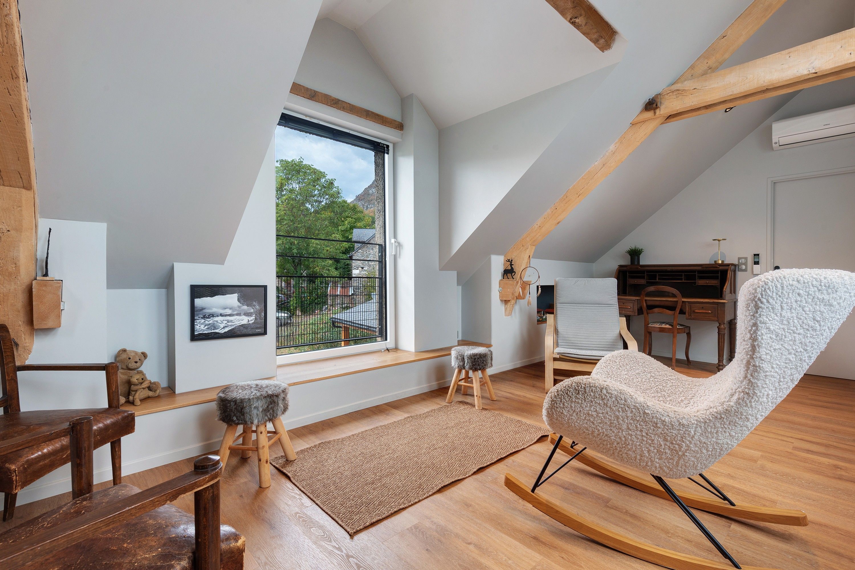 A loft-style room with a wooden floor, a rocking chair, a desk, and a large window overlooking a garden.