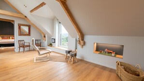 A spacious loft bedroom with a sloped ceiling, wooden beams, a large window, a bed, a chair, a small stool, and a basket. - Holiday Home “L’Escale Pibeste” with Mountain View, Wi-Fi and Air Conditioning (Agos-Vidalos)
