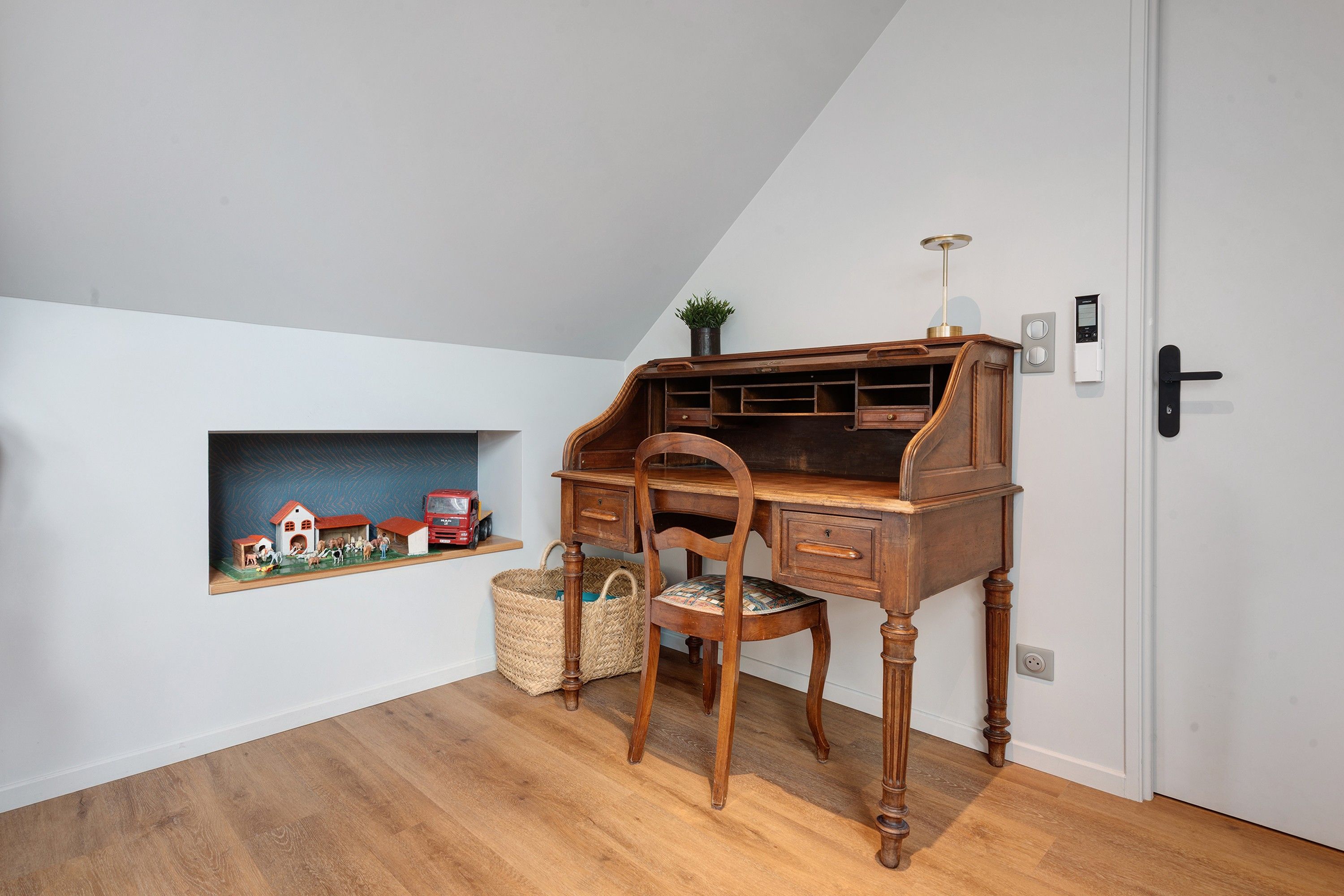 Une salle d’étude équipée d’un bureau en bois, d’une chaise, d’une petite étagère sur laquelle est posé un modèle de train miniature, et d’une porte dotée d’une serrure électronique.