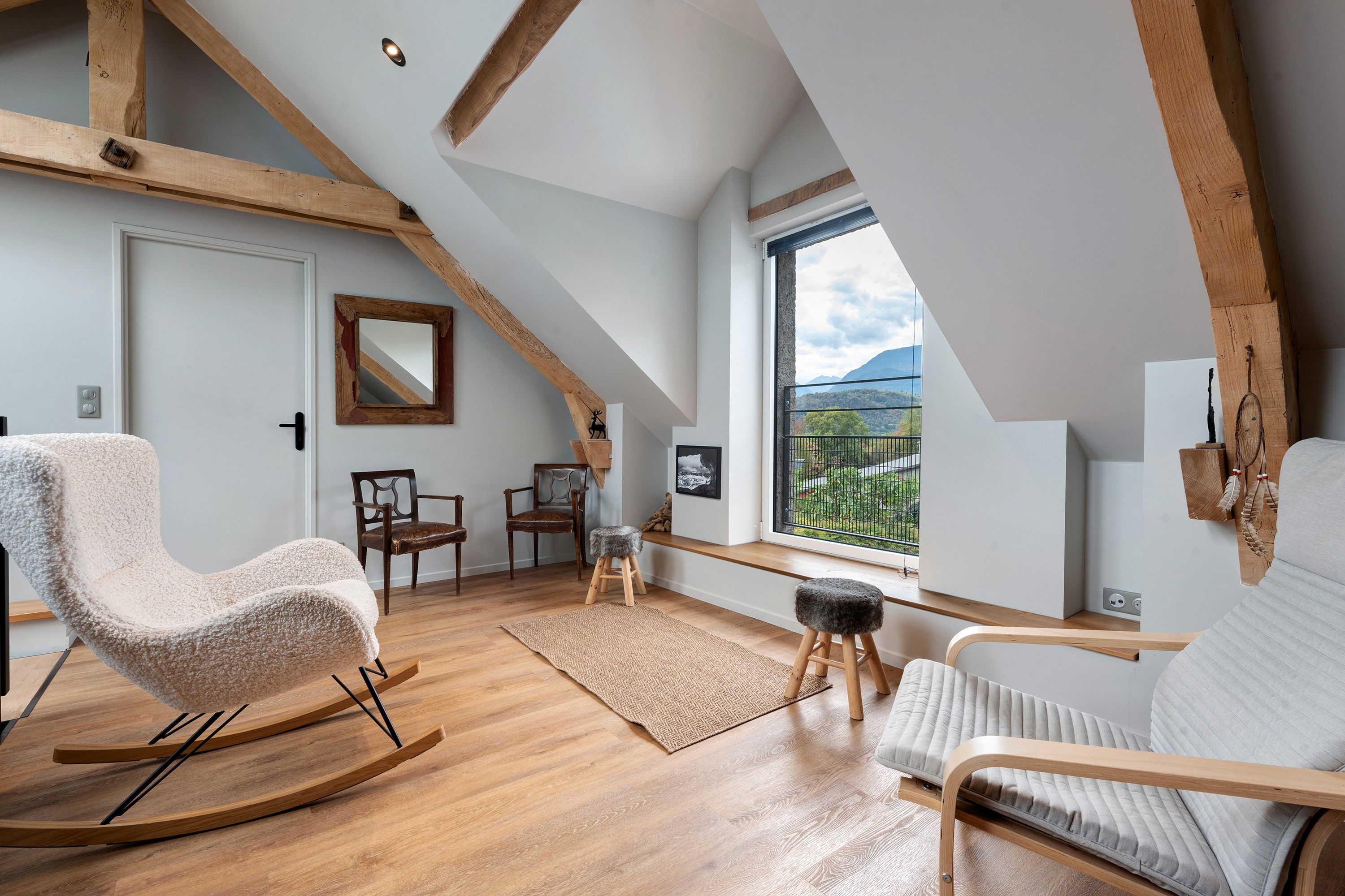 A loft-style room with exposed wooden beams, a large window with a view of greenery, a rocking chair, a cushioned bench, and a small stool.