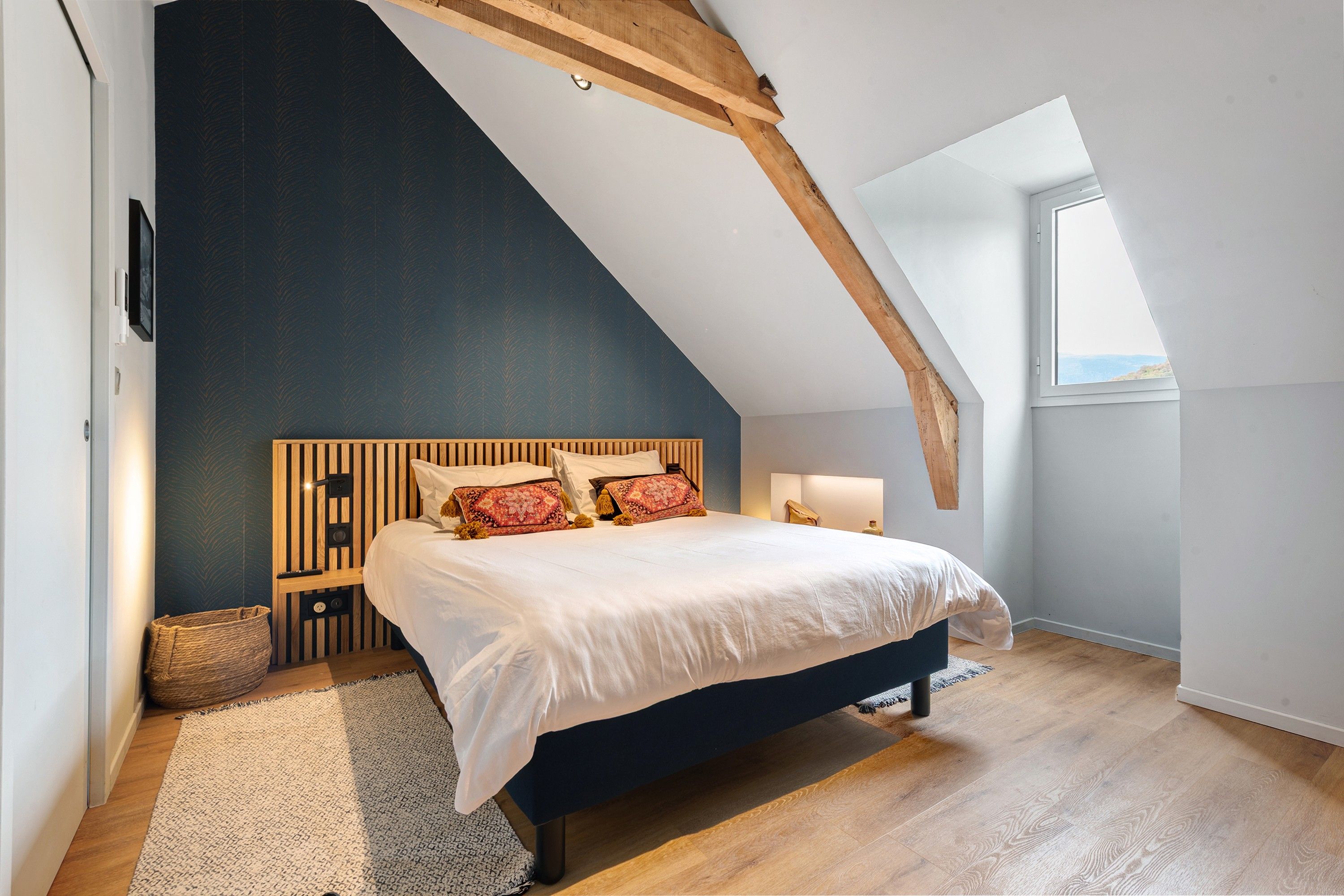 Une chambre avec un plafond en pente, un lit avec une literie blanche, une tête de lit en bois et une grande fenêtre.