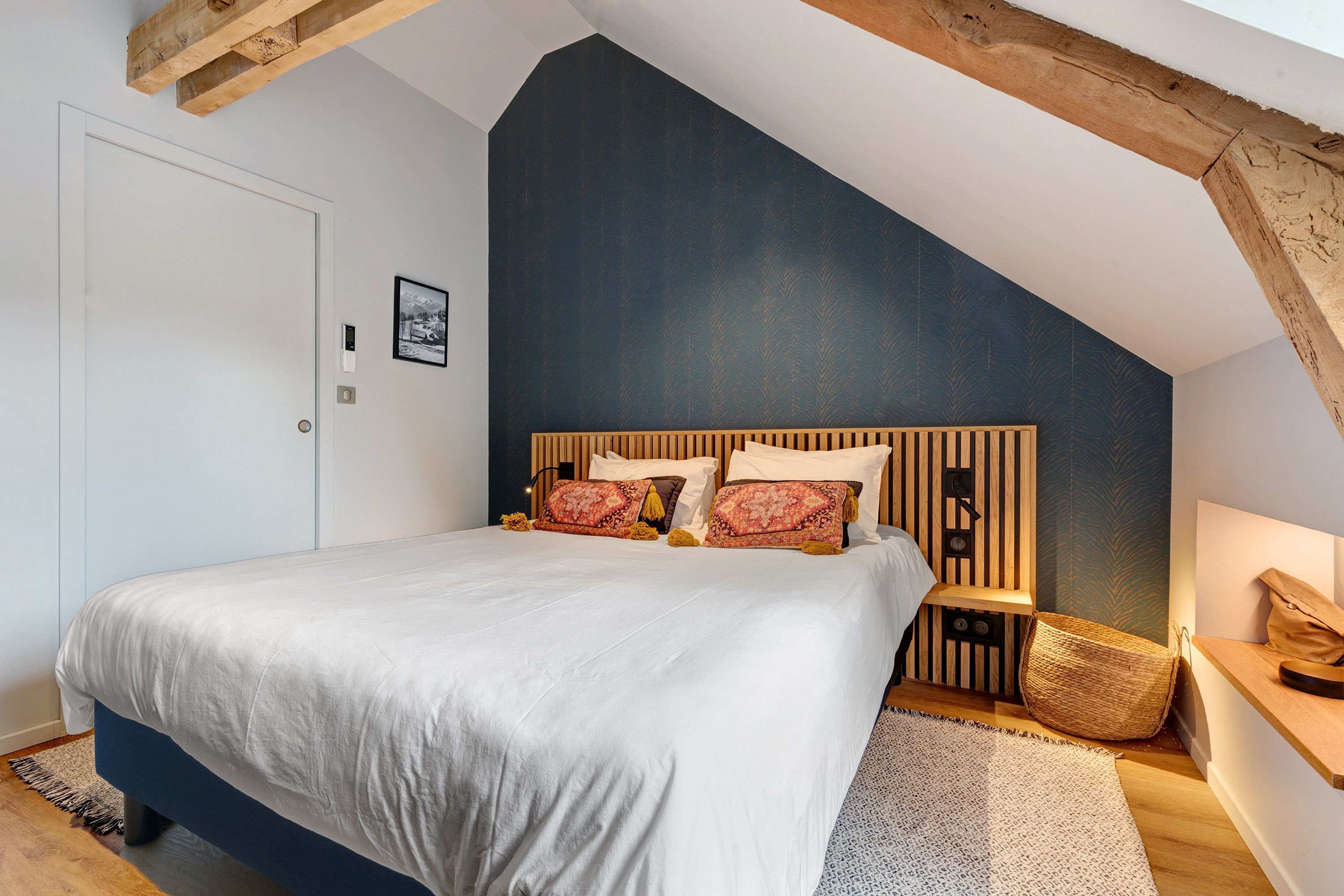 Un dormitorio con techo inclinado, cama con ropa de cama blanca, cabecero de madera y una pequeña mesita de noche con una lámpara.