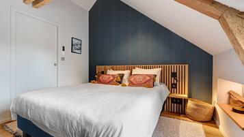Un dormitorio con techo inclinado, cama con ropa de cama blanca, cabecero de madera y una pequeña mesita de noche con una lámpara.