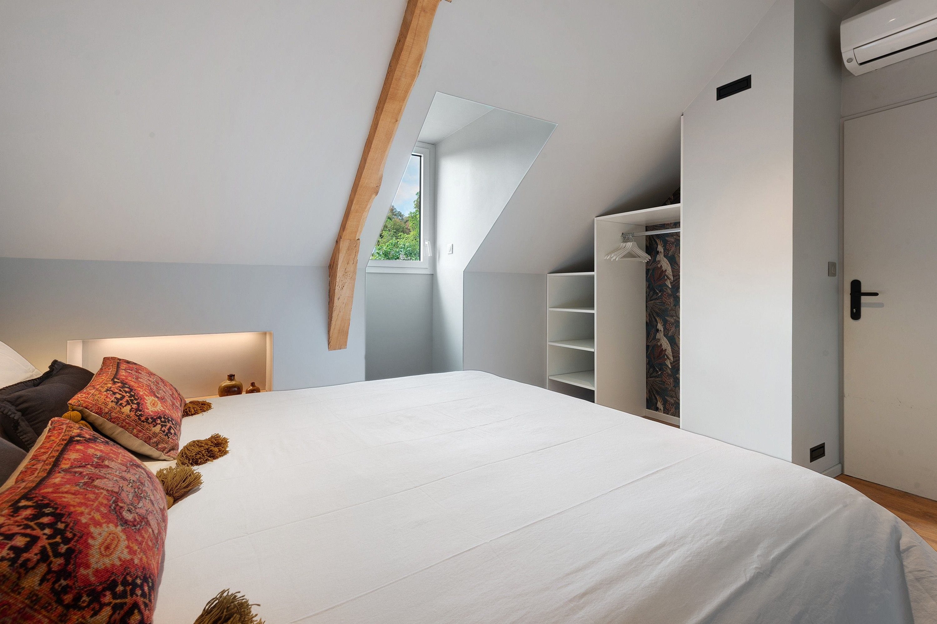 A bedroom with a sloped ceiling, a bed with patterned pillows, a wardrobe, and a window with a view.